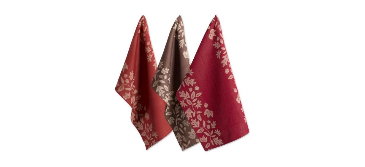 A set of 3 dish towels: one is orange, one is brown and one is red. All have a beige design on the edges of fall leaves